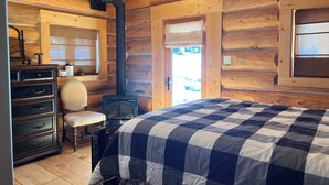 A log cabin bedroom with a bed, dresser, chair, and a wood-burning stove.