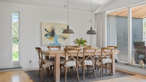 A dining area with a wooden table and chairs, a painting on the wall, and a view through large windows.