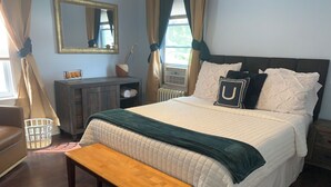 Quarto com cama, mesa de cabeceira, banqueta, cômoda, espelho e cortinas.