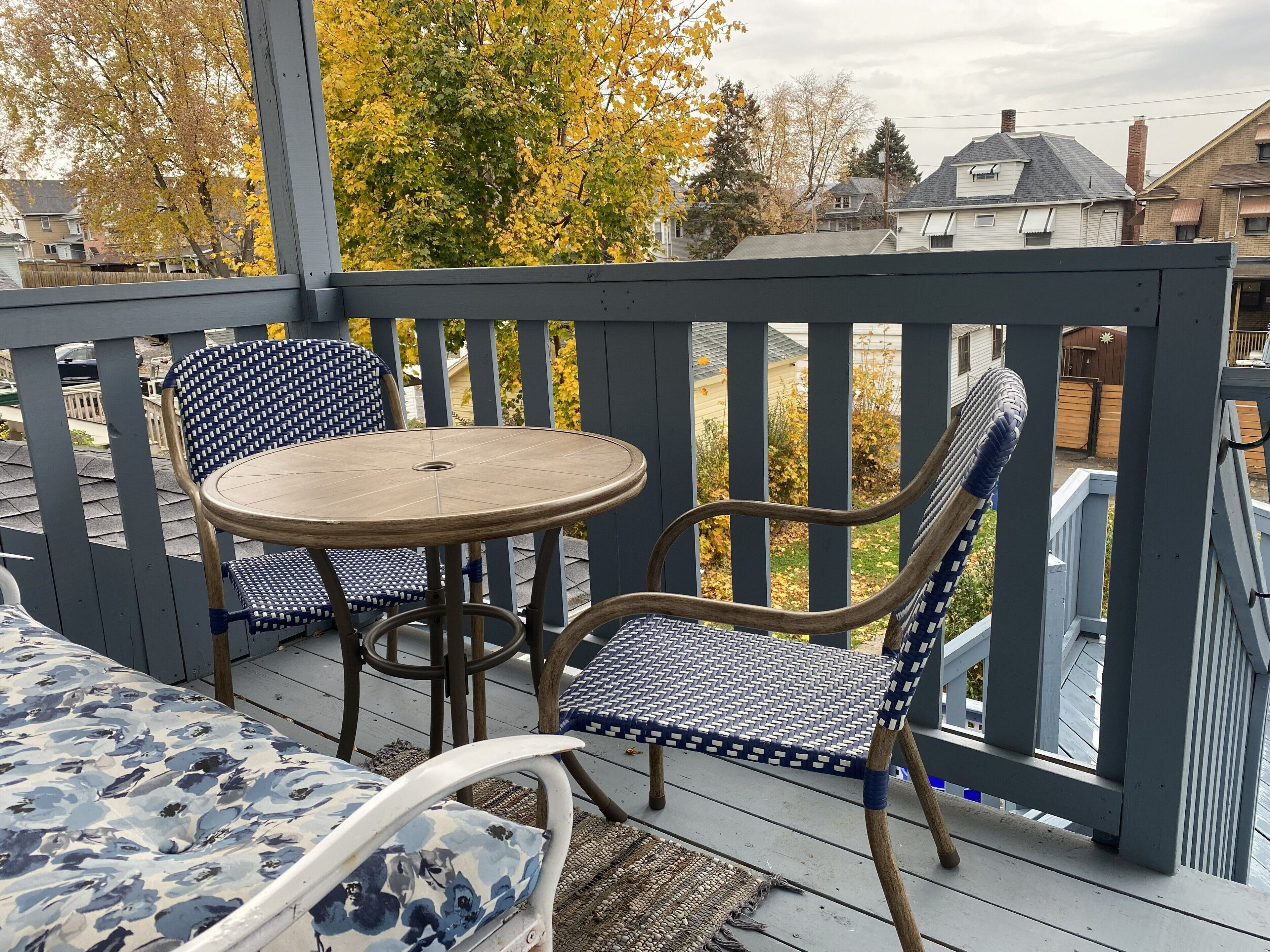 Ett takförsett utomhusområde med sittplatser, ett runt bord och två mönstrade stolar, med utsikt över ett bostadsområde.