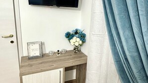 Une pièce avec un téléviseur fixé au mur, une coiffeuse en bois avec un miroir, un petit vase avec des fleurs et une porte avec une poignée.