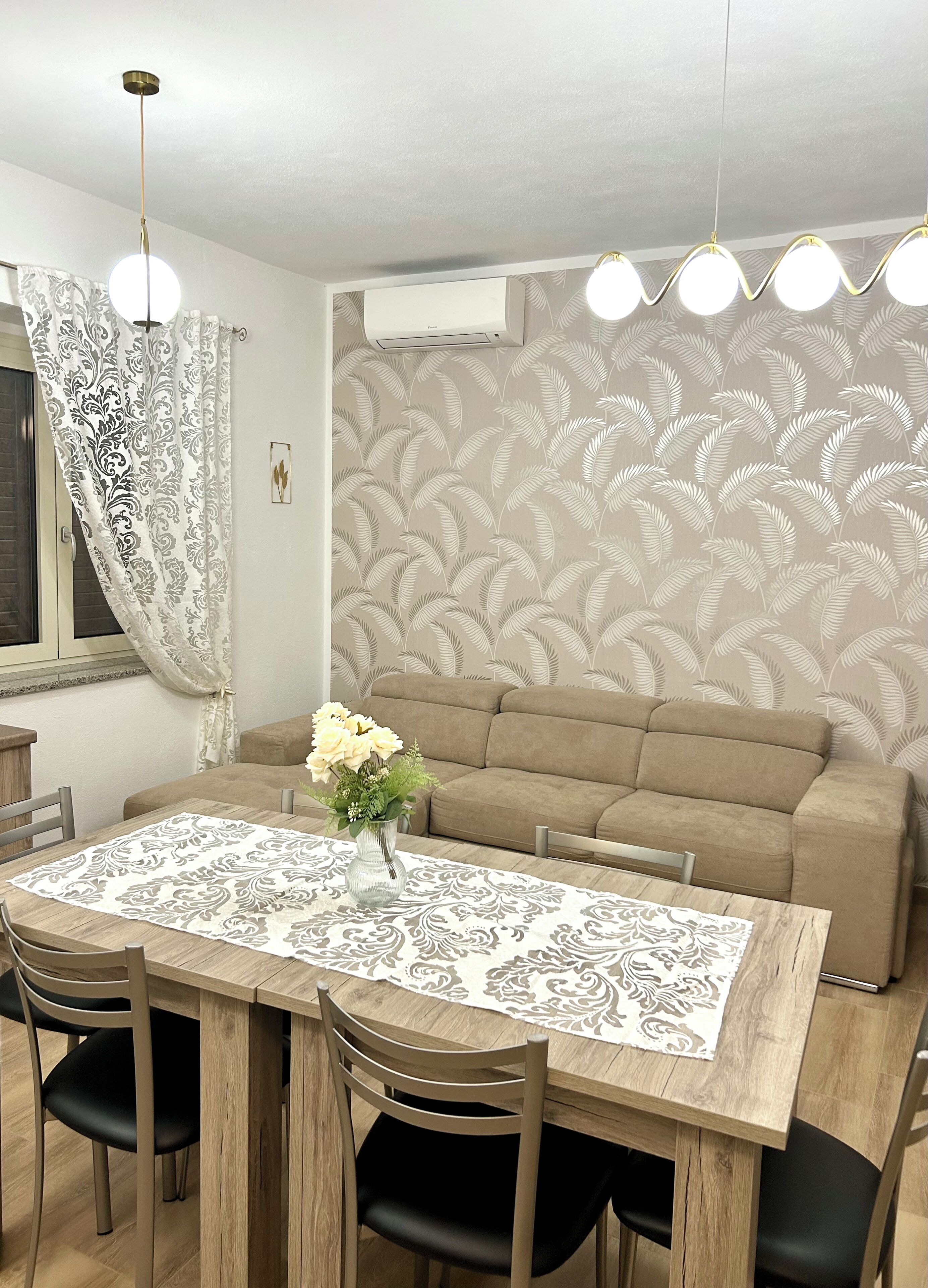 Un coin repas avec une table en bois, des chaises noires, un vase de fleurs et un mur orné d’un motif.