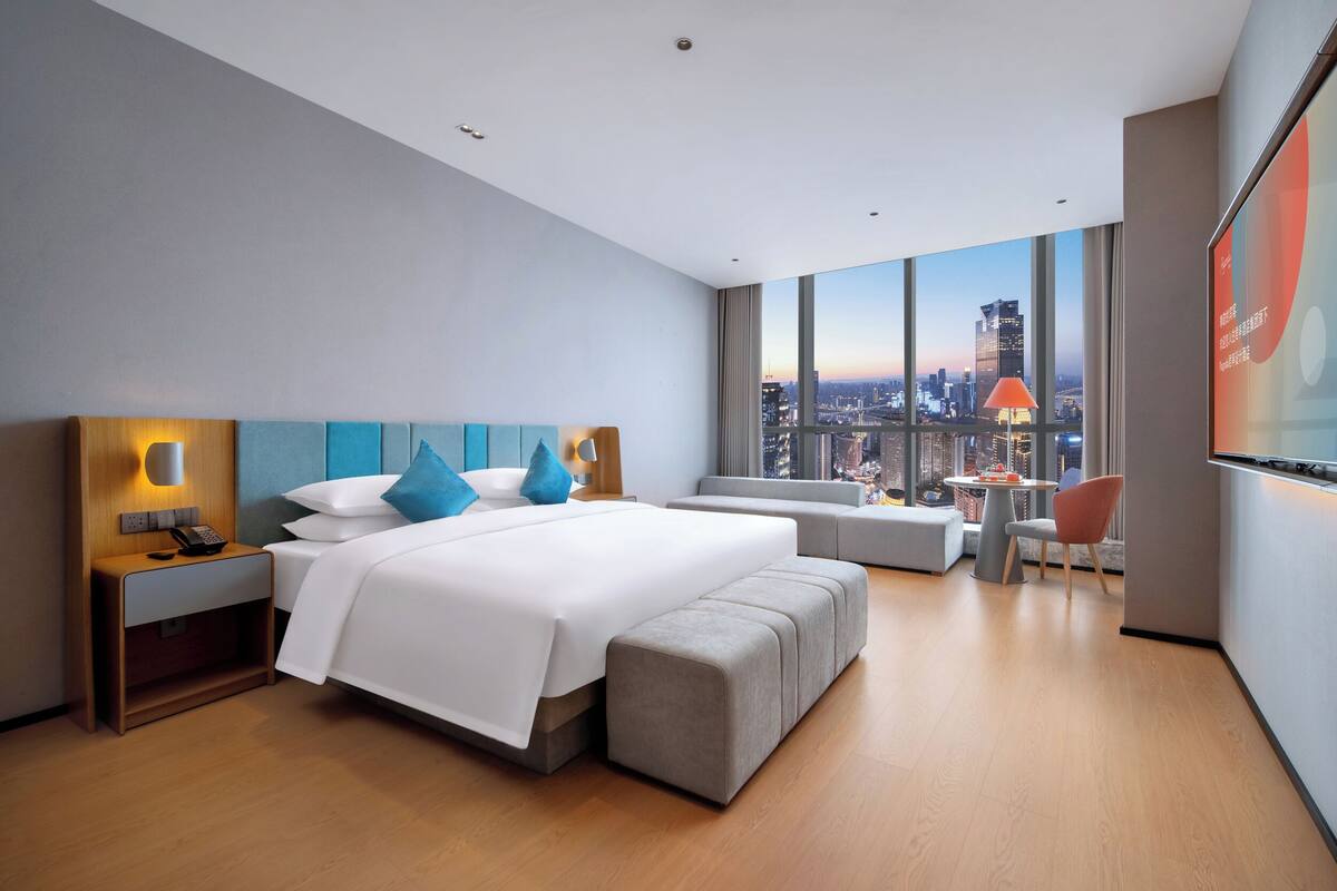 A modern hotel room with a large bed, a seating area, and a city view.