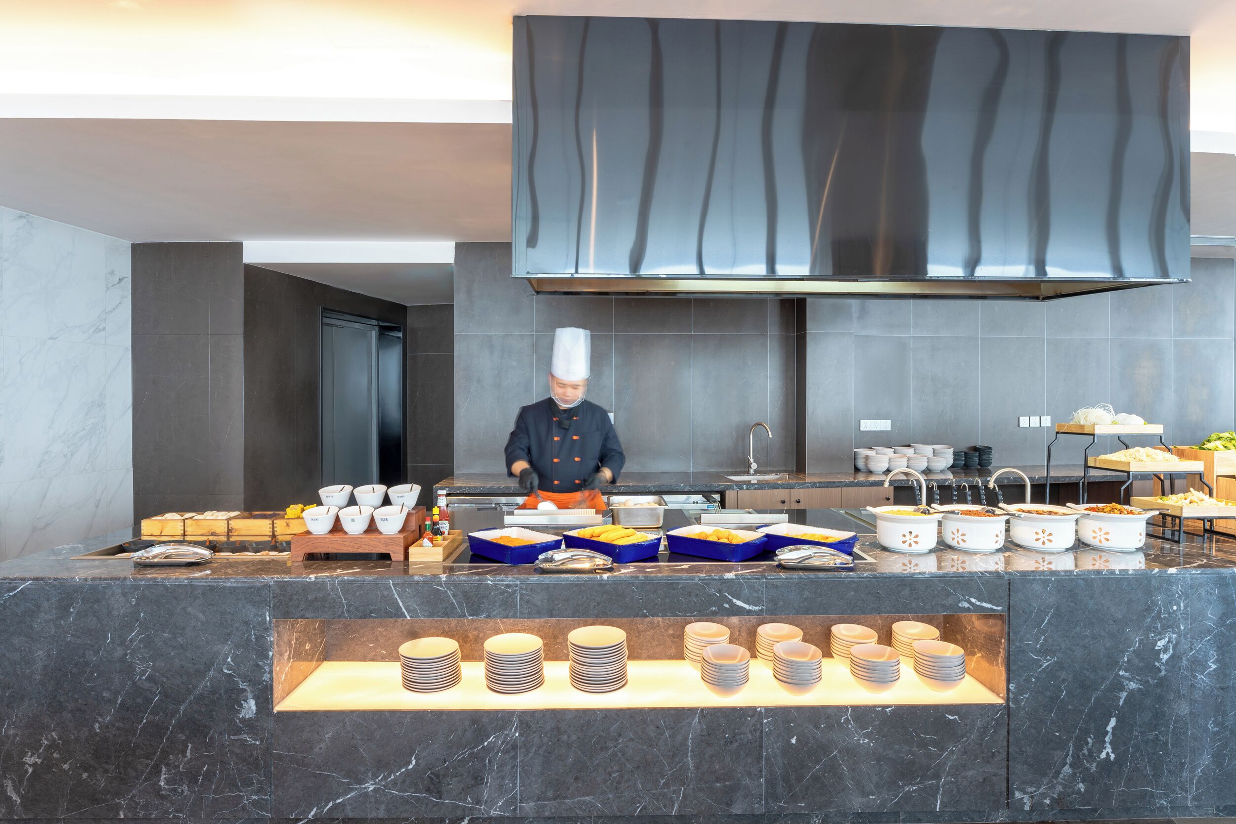 Una cocina moderna con un chef preparando comida, estaciones de buffet con alimentos y platos y tazones ordenados.
