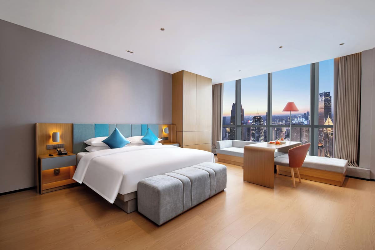 A modern hotel room with a large bed, a desk, and a view of a cityscape.