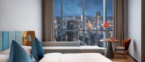 Una habitación de hotel moderna con un amplio ventanal que ofrece vista a la ciudad, una cama con almohadas azules, una mesita de noche con una lámpara y una silla.