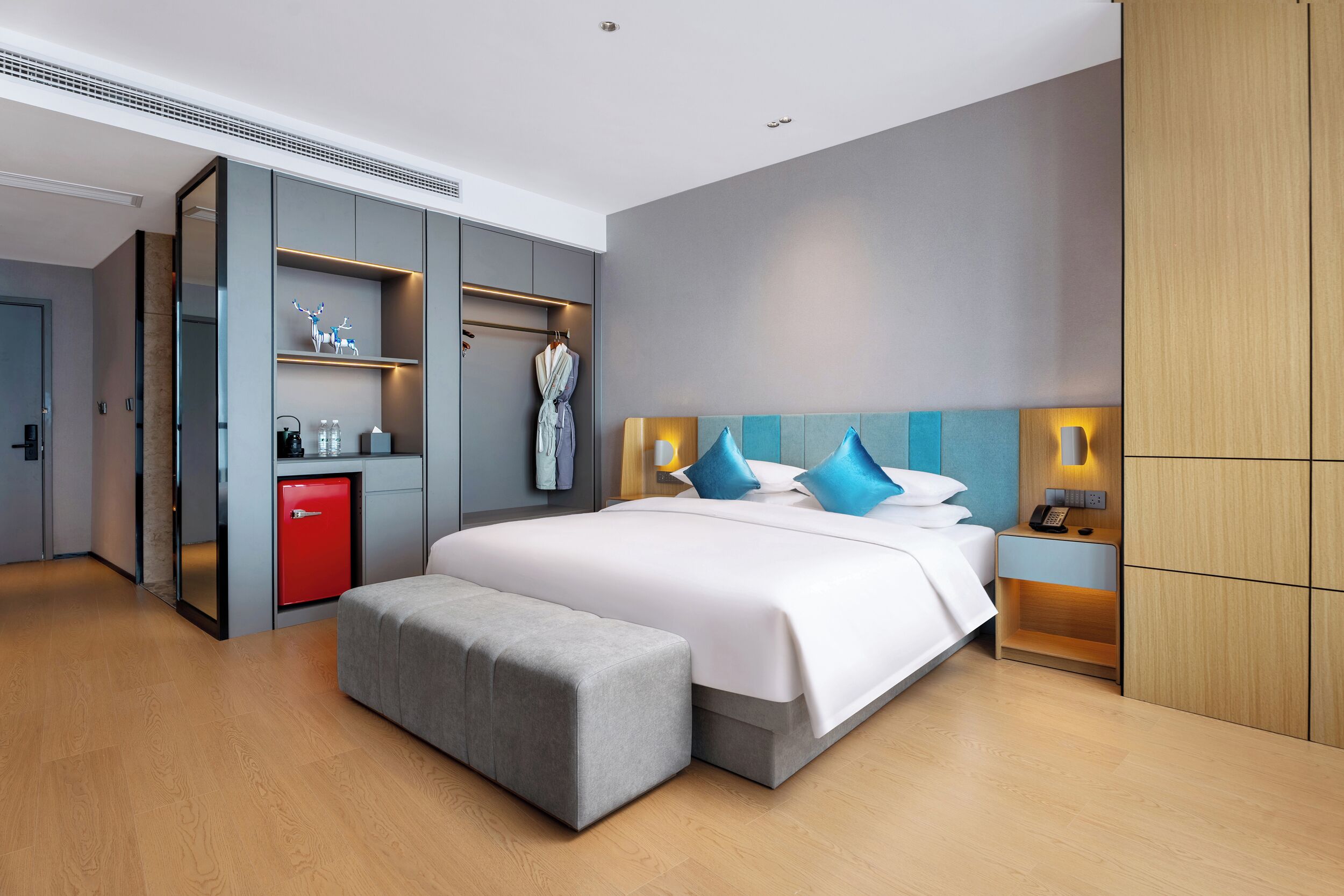 Una habitación de hotel moderna con una cama grande, un banco gris, un minibar rojo y un armario con espejo.