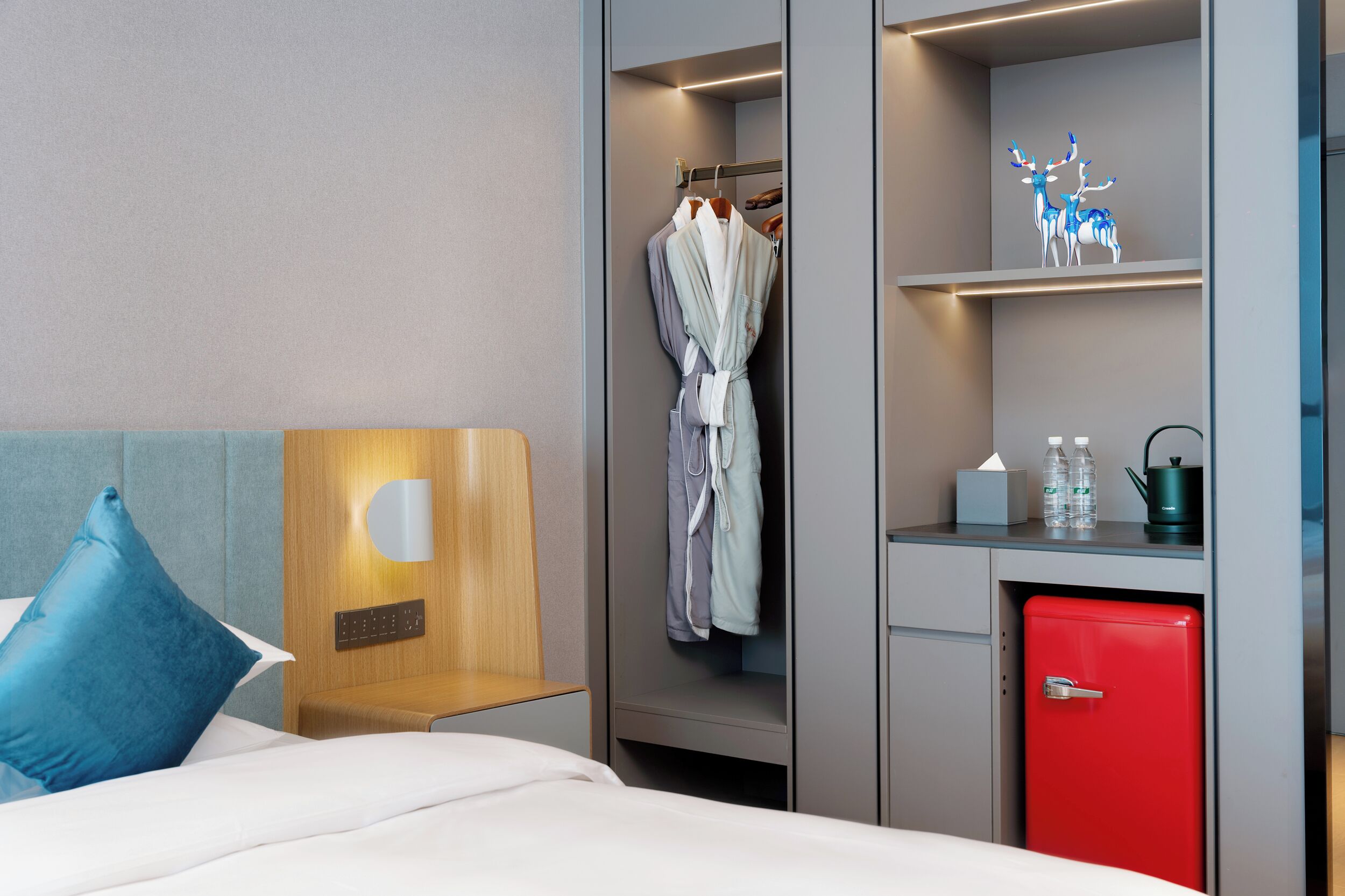 Une chambre d’hôtel comprenant un lit, une tête de lit en bois, une armoire avec un peignoir, un mini-réfrigérateur rouge et une étagère avec des objets décoratifs.