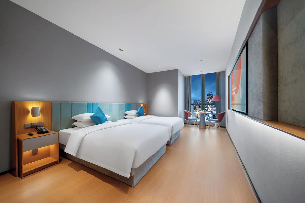 A modern hotel room with a large bed, bedside tables, a desk, and a view of the cityscape.