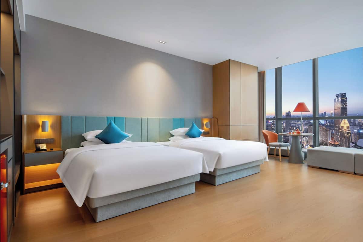 A modern hotel room with two beds, a city view, a small table, and a chair.