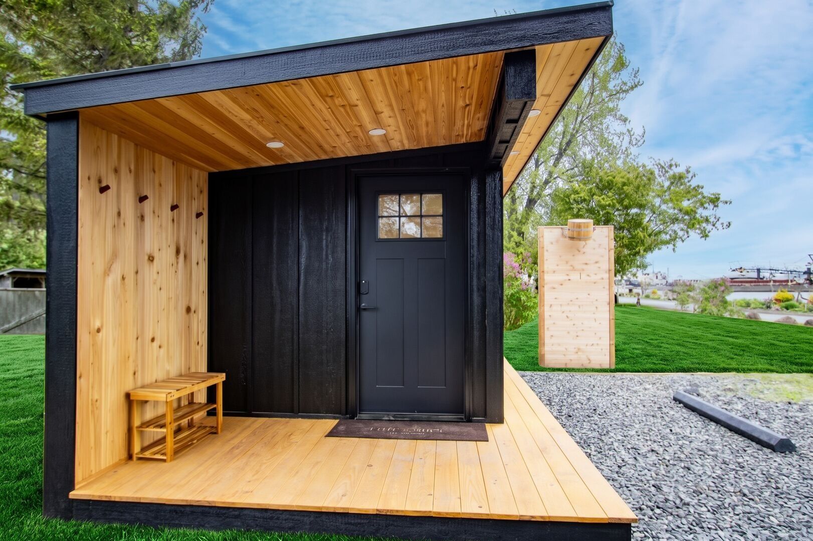 A modern, compact building with a wooden exterior and a black door, featuring a small bench and a doormat in front.