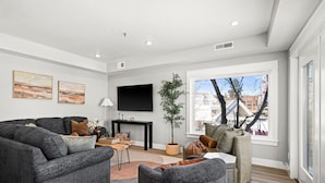A modern living room with a grey sofa set, a coffee table, and a TV mounted on the wall. There is a large window providing a view of the outside. - Luxury Condo w/ Beach Access | Dragestil 717-2 (Duluth)