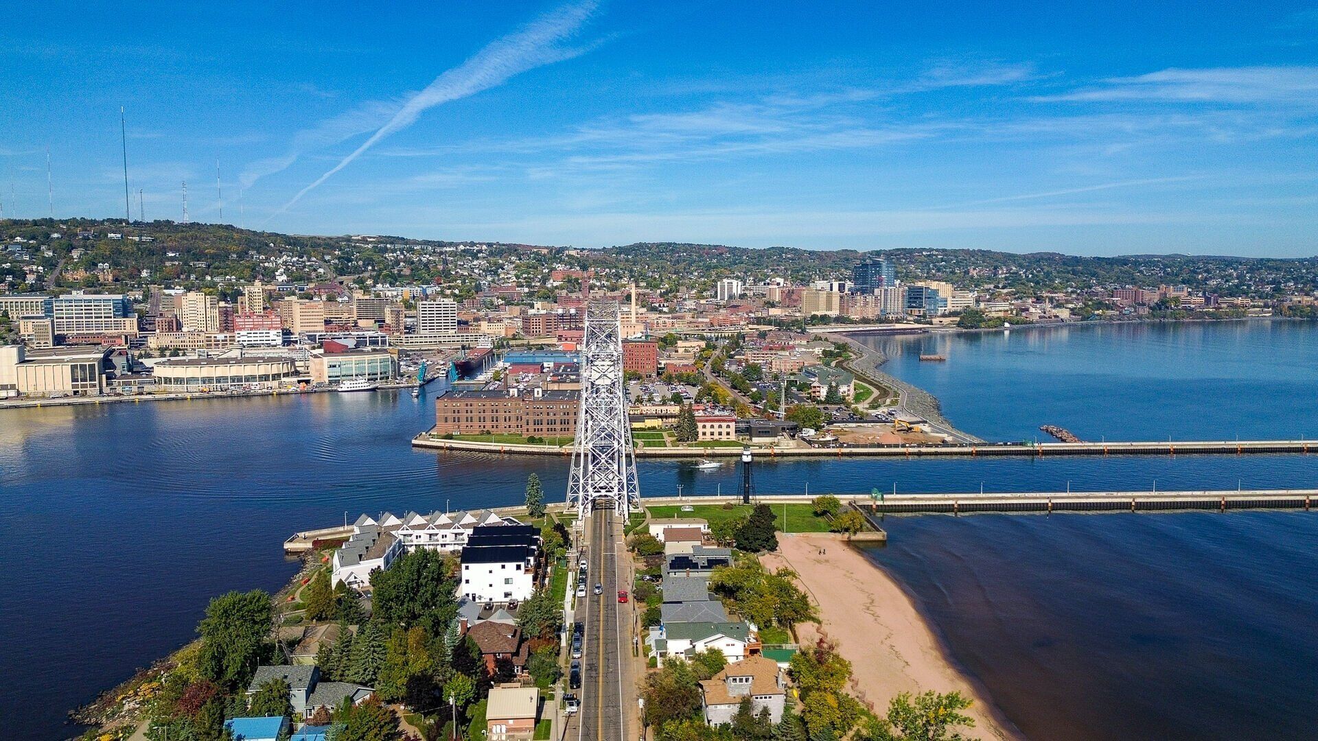 Un paysage urbain côtier avec un pont imposant, des bâtiments situés au bord de l’eau et une chaîne de collines en arrière-plan.