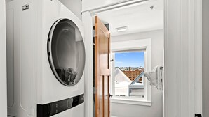 A laundry room with stacked washers and dryers, a wooden door leading to a room with a window and a view of buildings. - Penthouse Condo w/ Beach Access | Dragestil 717-3 (Duluth)