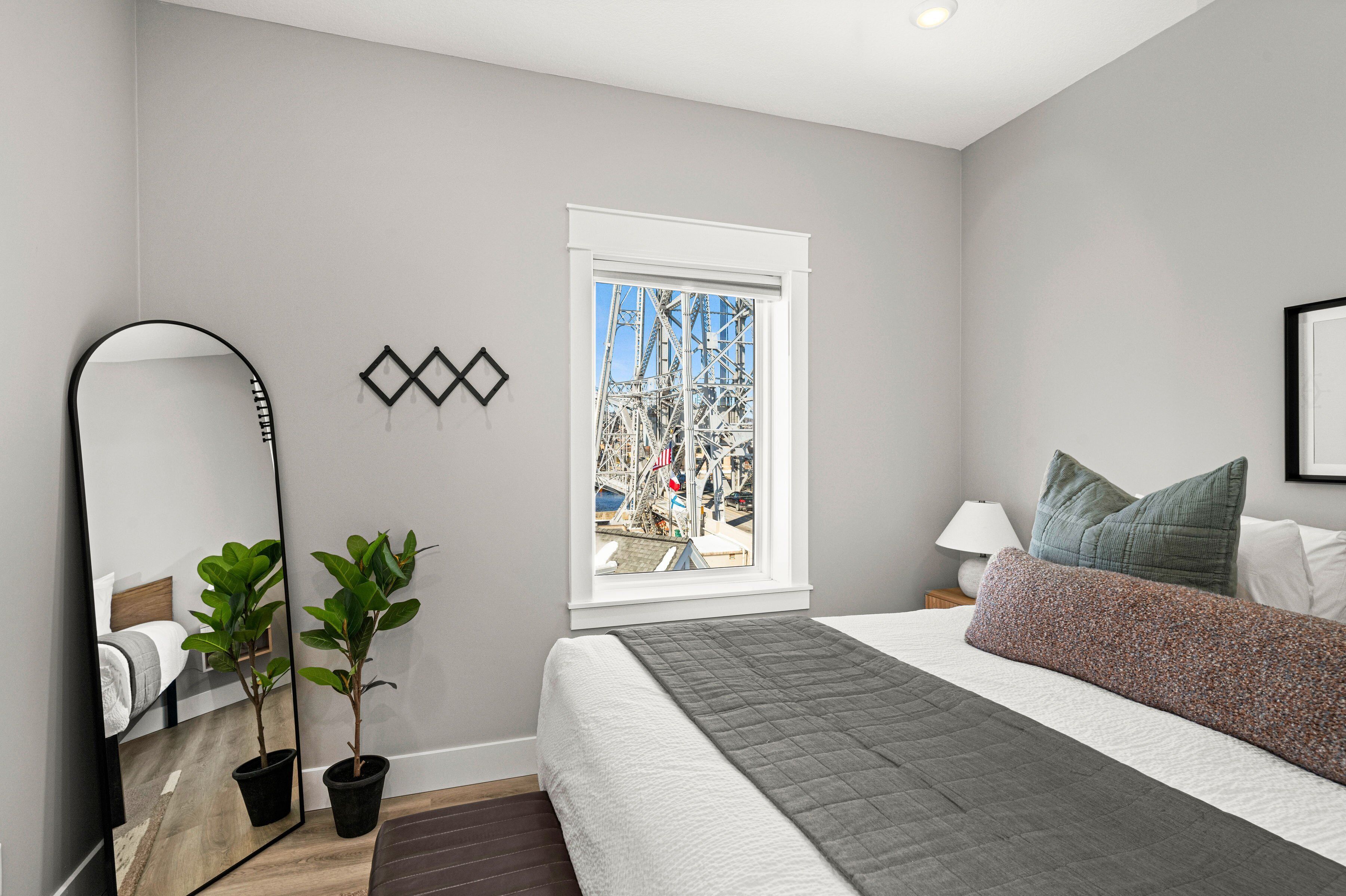A bedroom with a bed, a window with a view of a construction site, a mirror, and two potted plants.