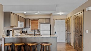 A kitchen with wooden cabinets, a stainless steel refrigerator, a microwave, and a coffee maker. There are three wooden stools at the counter.