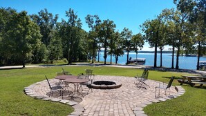 A circular outdoor seating area with a fire pit, surrounded by a paved walkway and grassy areas, overlooking a lake with a dock and pier.