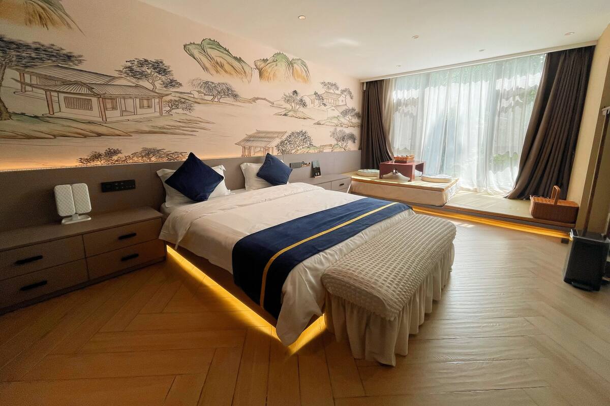 Una habitació d'hotel amb un llit gran, una taula de nit, cortines a la finestra i un mural a la paret.