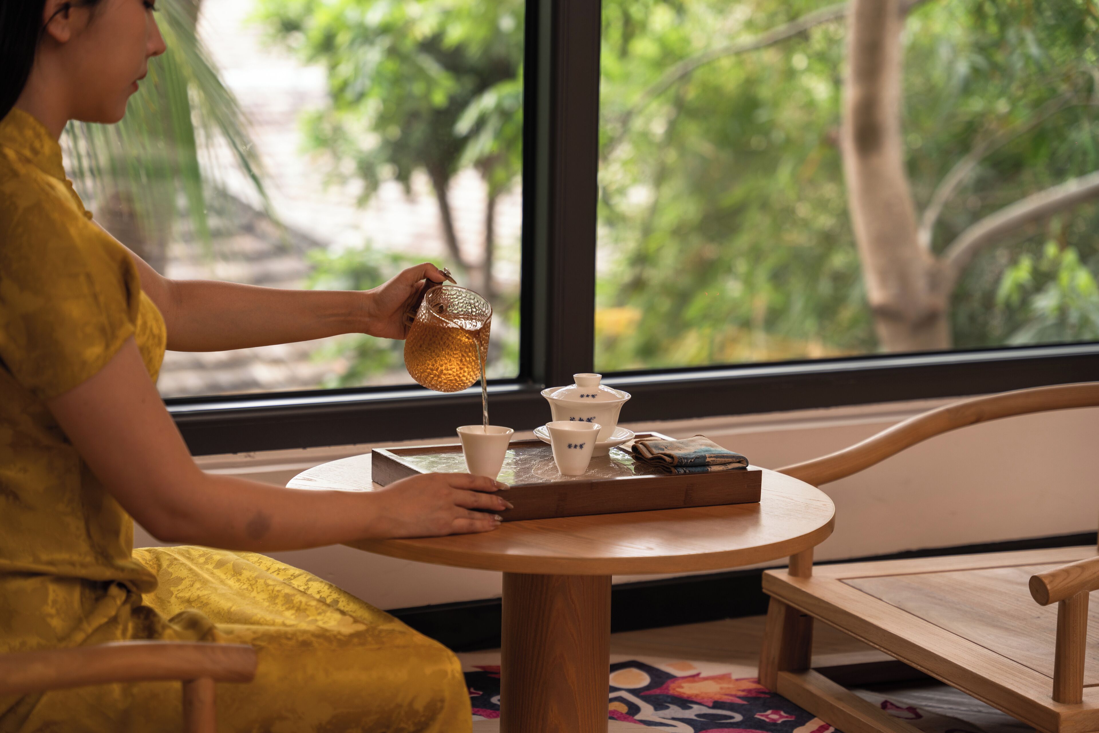 A person pouring a drink into a cup at a wooden table with a tray of teaware.