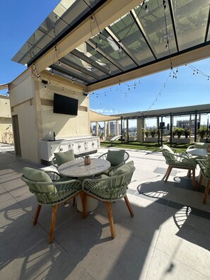 Une terrasse extérieure avec une table ronde et des chaises, un barbecue encastré et un écran de télévision.