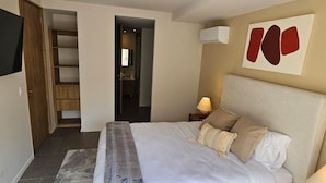 A bedroom with a bed, a nightstand, a lamp, a wall-mounted air conditioner, and a door leading to a bathroom.