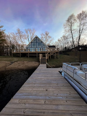 A wooden dock leading to a house with a balcony, surrounded by trees and a body of water.