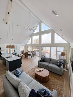 A spacious living area with a high ceiling, large windows, a dining area, and a kitchen.