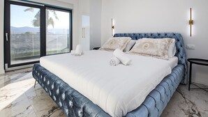 A large bed with a tufted headboard, a marble floor, and a view of palm trees through a sliding glass door.