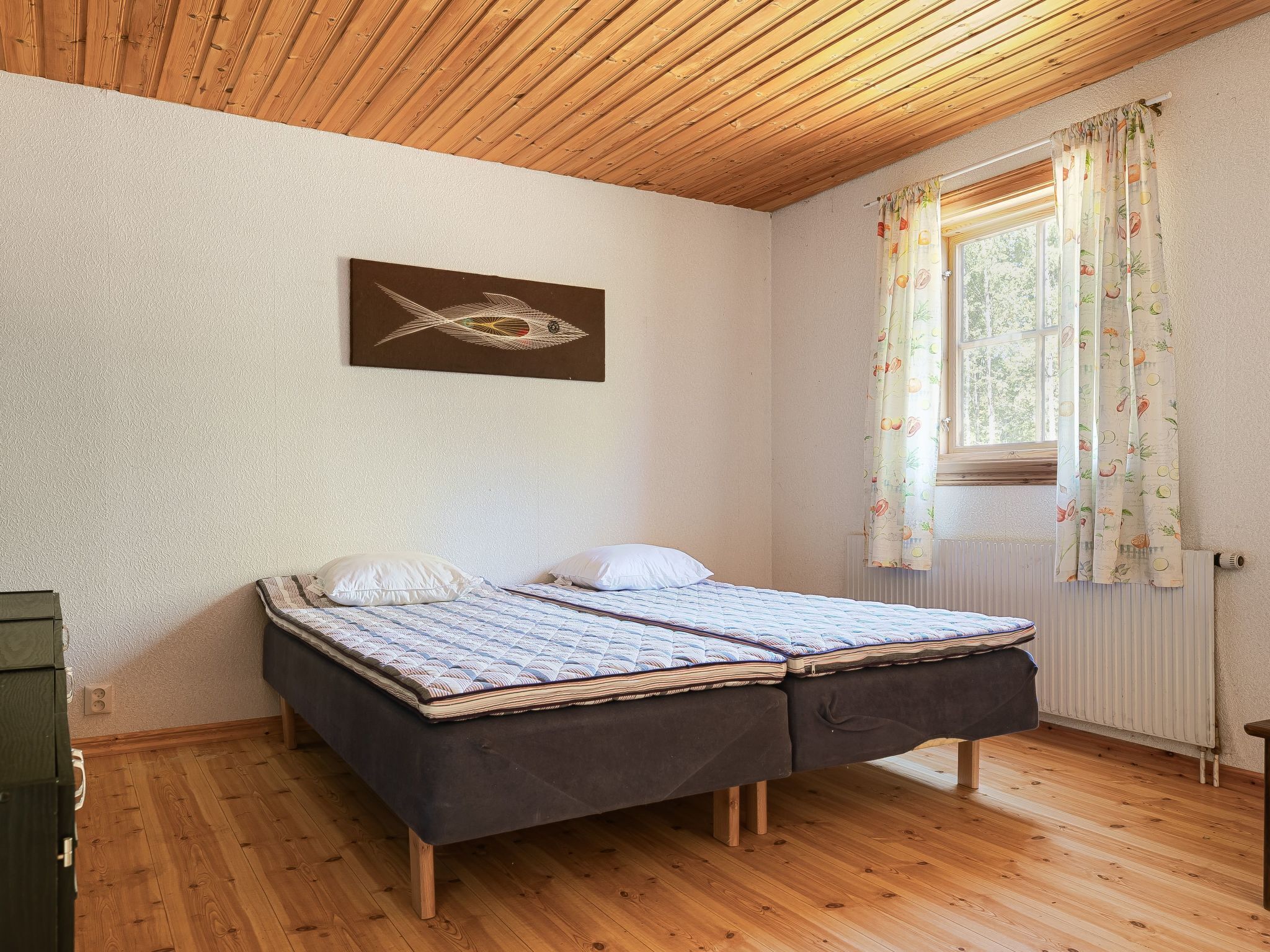 Ein Schlafzimmer mit Holzbalkendecke, einem Bett mit Tagesdecke, einem an der Wand hängenden Fischgemälde und einem Fenster mit Vorhängen.