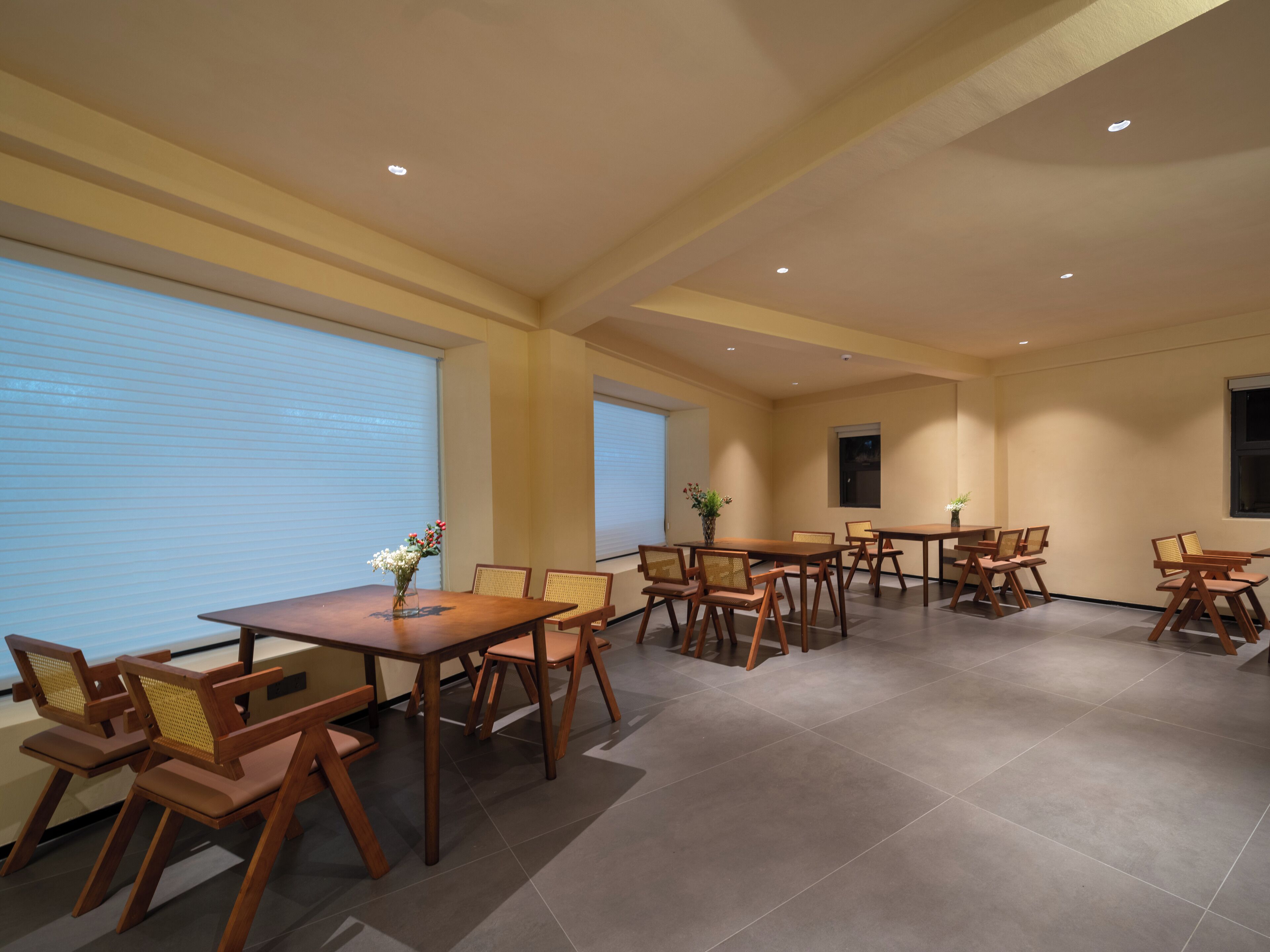 A spacious dining area with multiple tables and chairs, large windows with blinds, and recessed ceiling lighting.