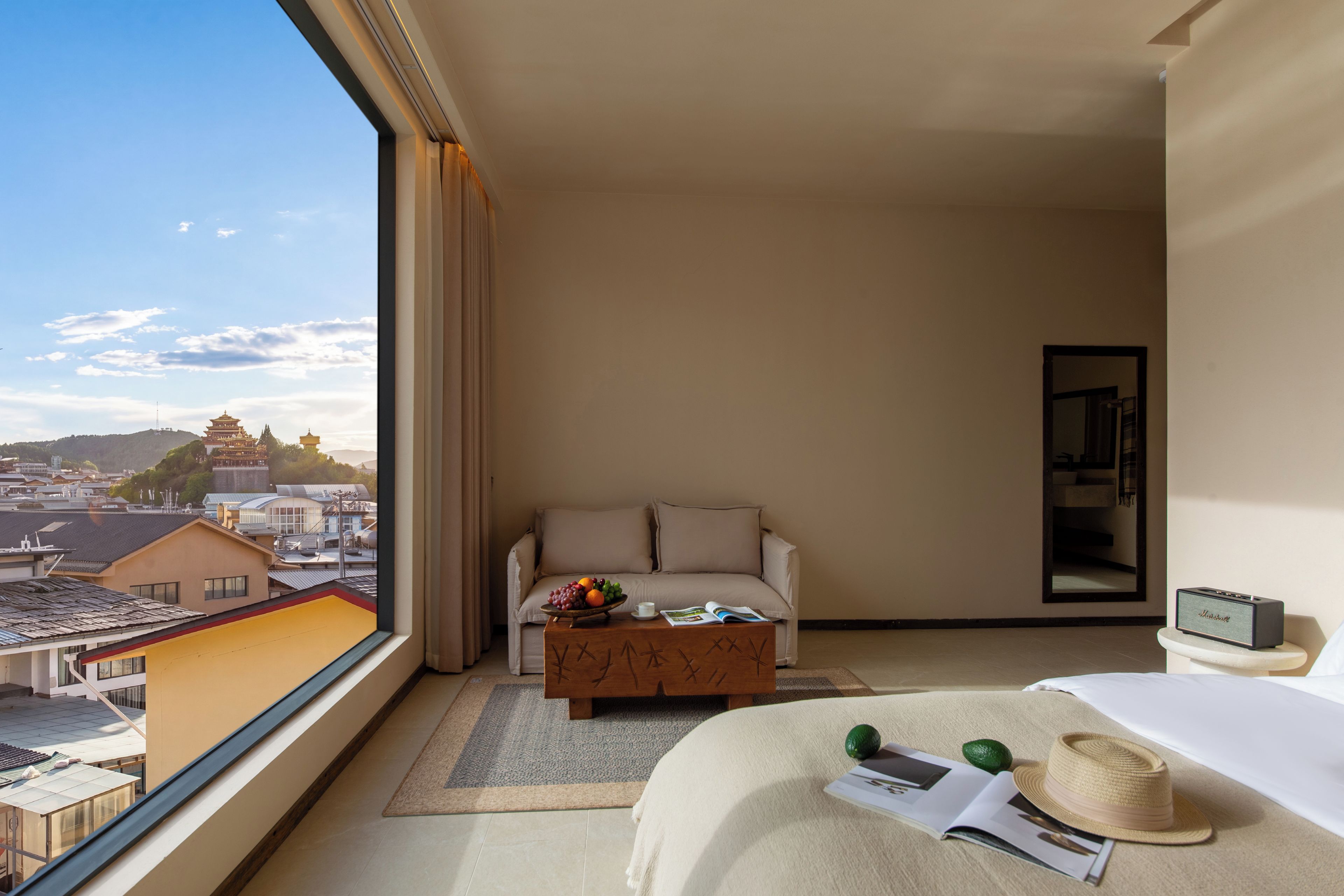 A hotel room with a large window, a bed, a sofa, a coffee table with fruit, and a small table with a book and a hat.