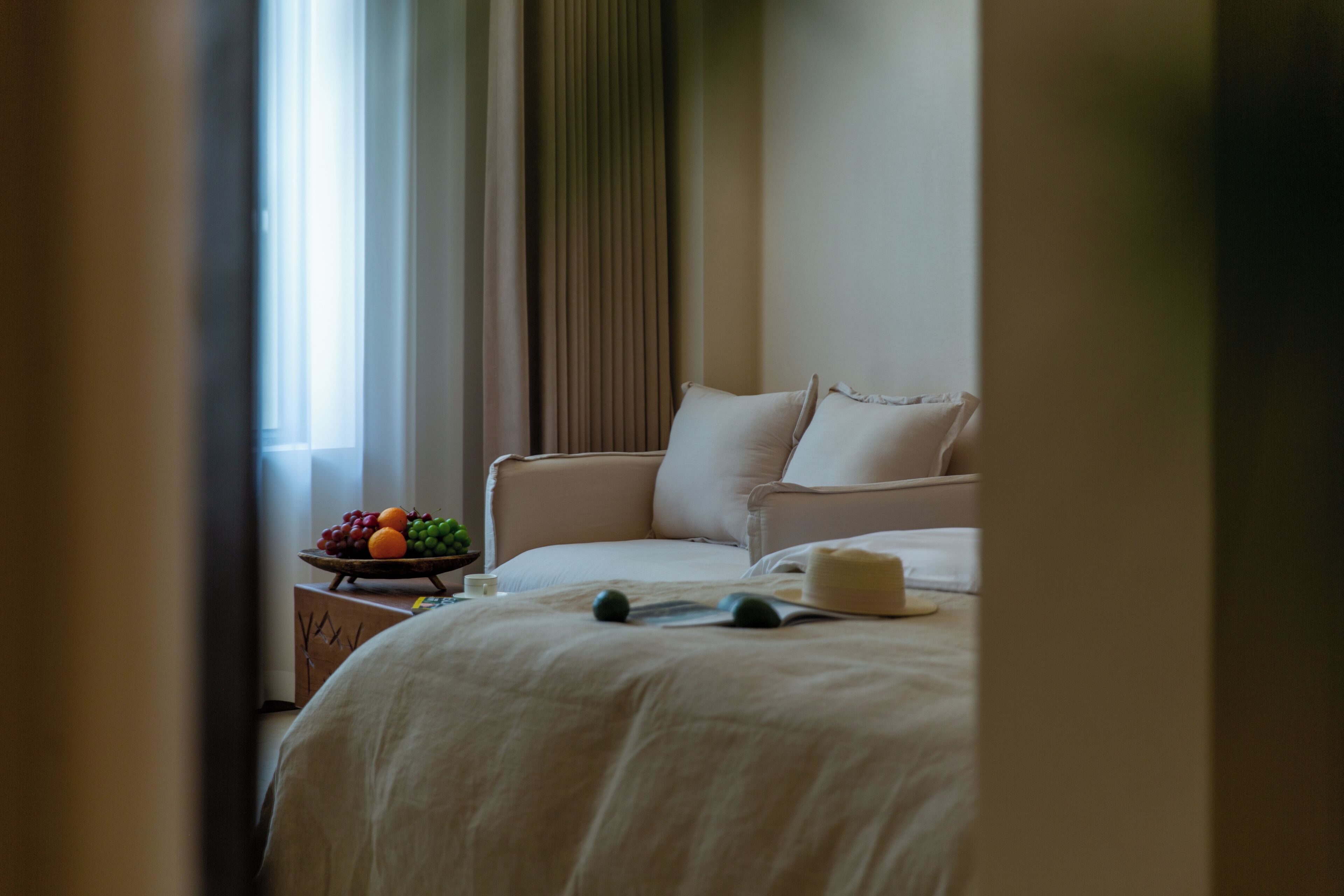 A hotel room with a bed, pillows, a bedside table with a fruit bowl, and a window with curtains.