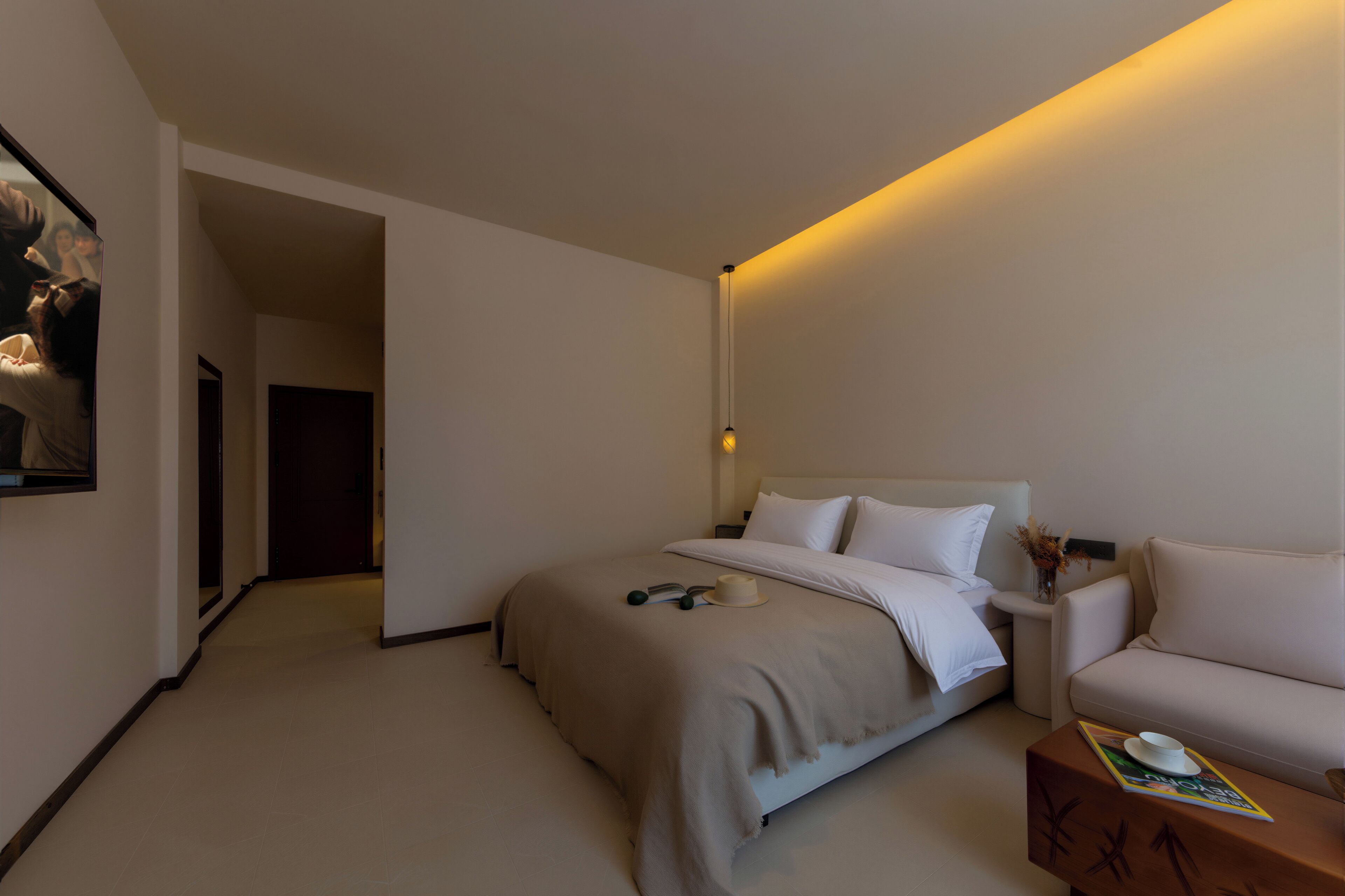 A hotel room with a bed, a nightstand, a bedside table with a vase, and a door leading to another room.