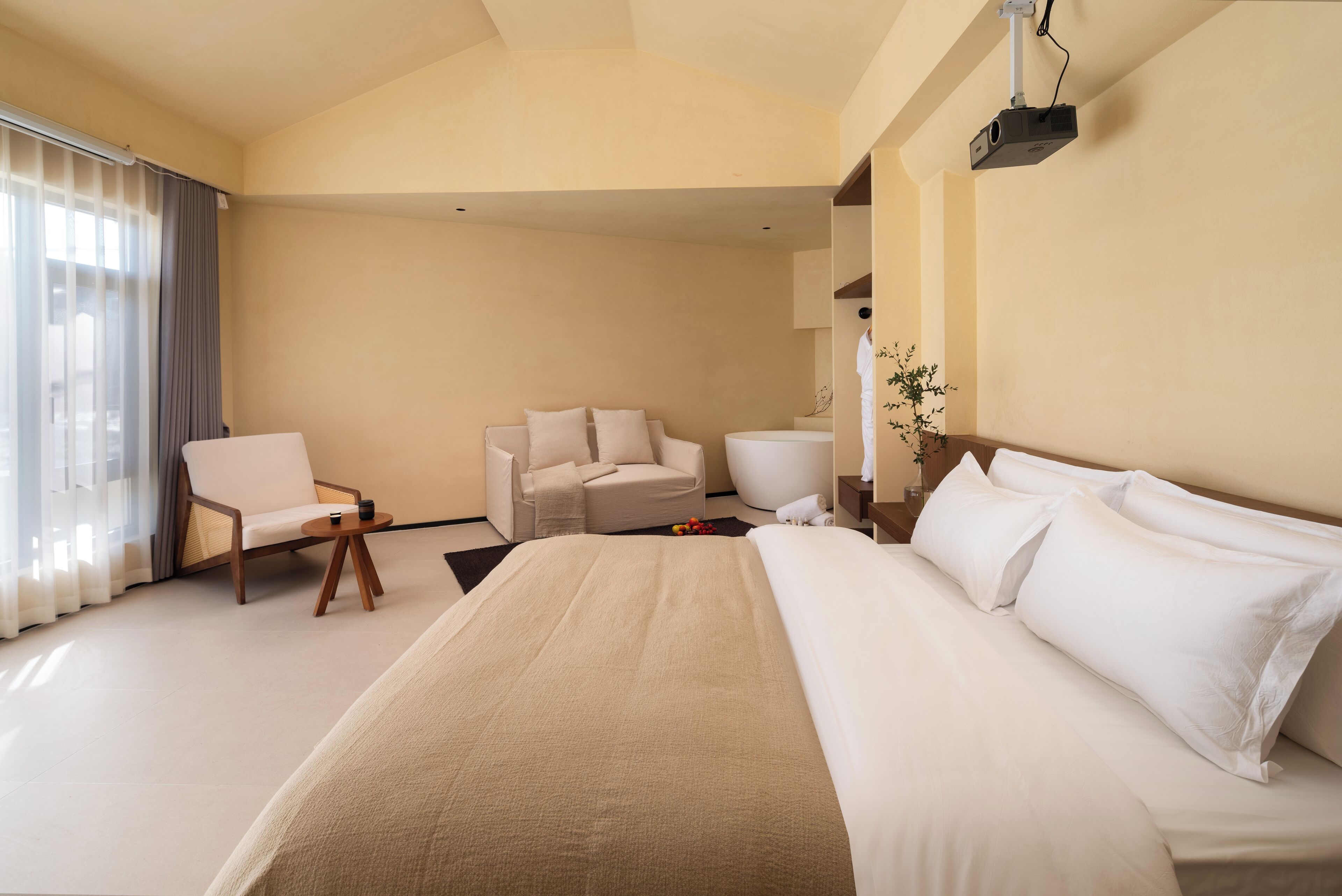 A modern hotel room with a large bed, a sofa, a small table, and a projector mounted on the ceiling.