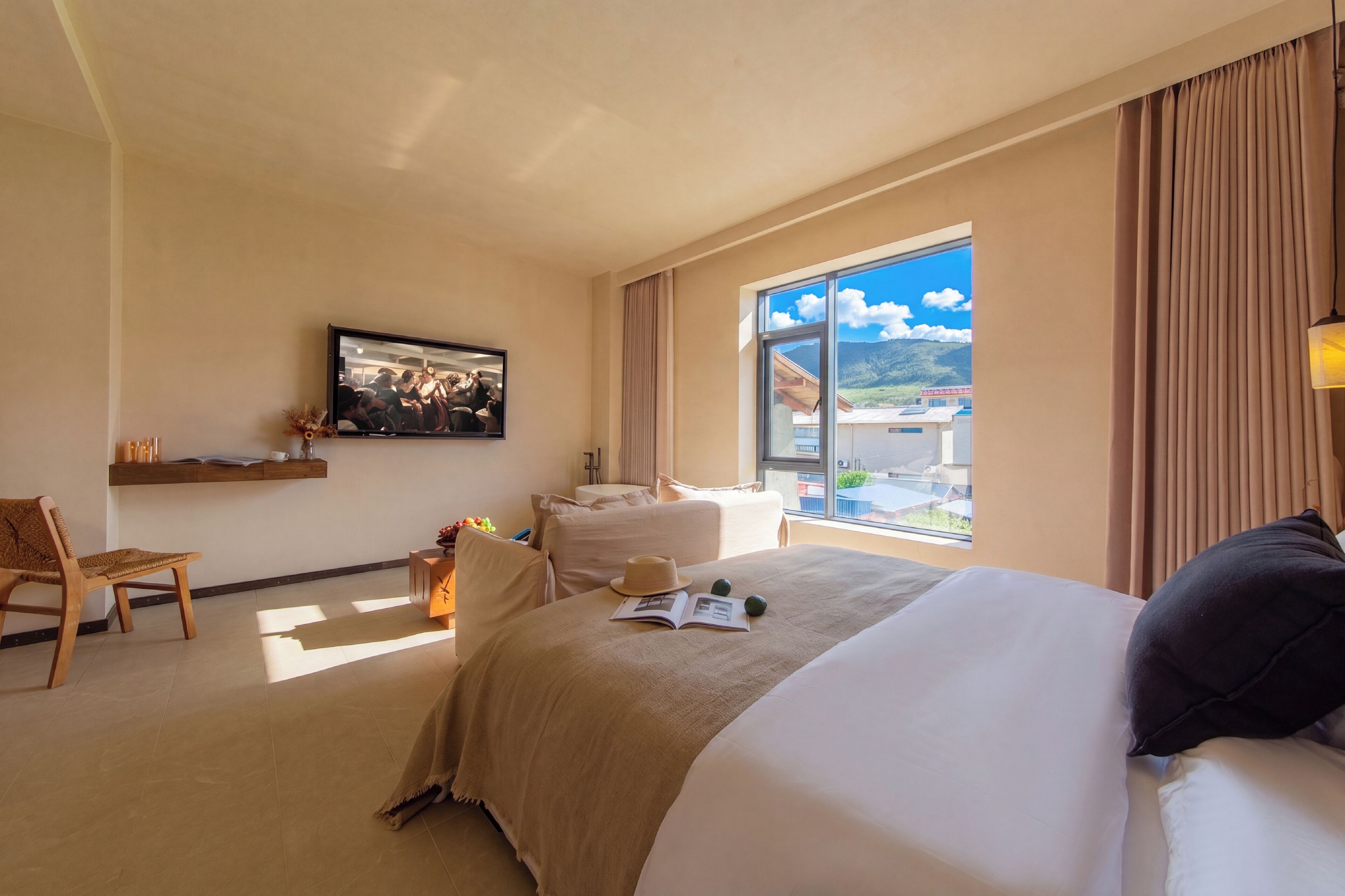 A hotel room with a bed, a chair, a desk, a television, and a window with a view of buildings and mountains.