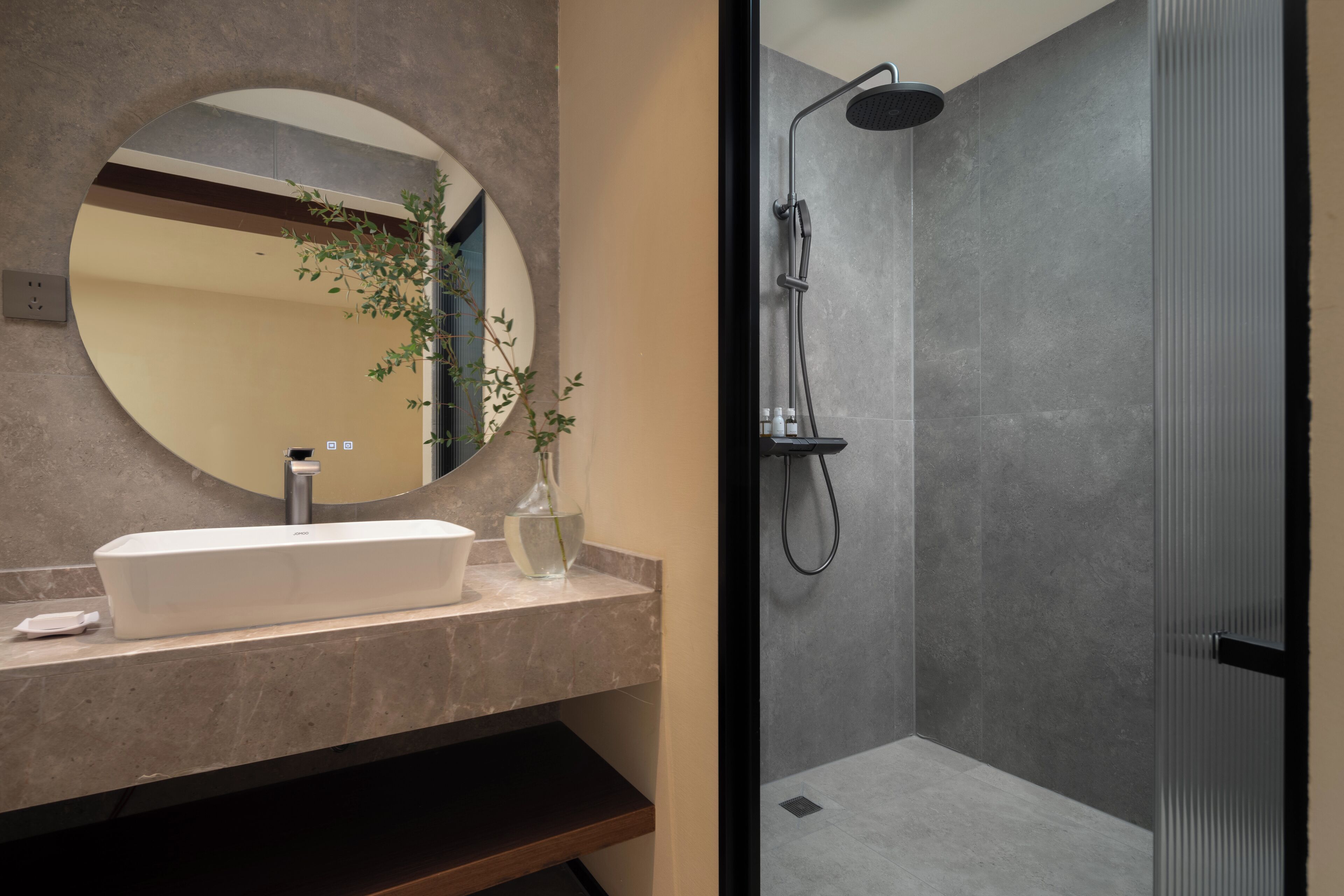 Ein Badezimmer mit rundem Spiegel, Waschbecken, Dusche und einer Pflanze.