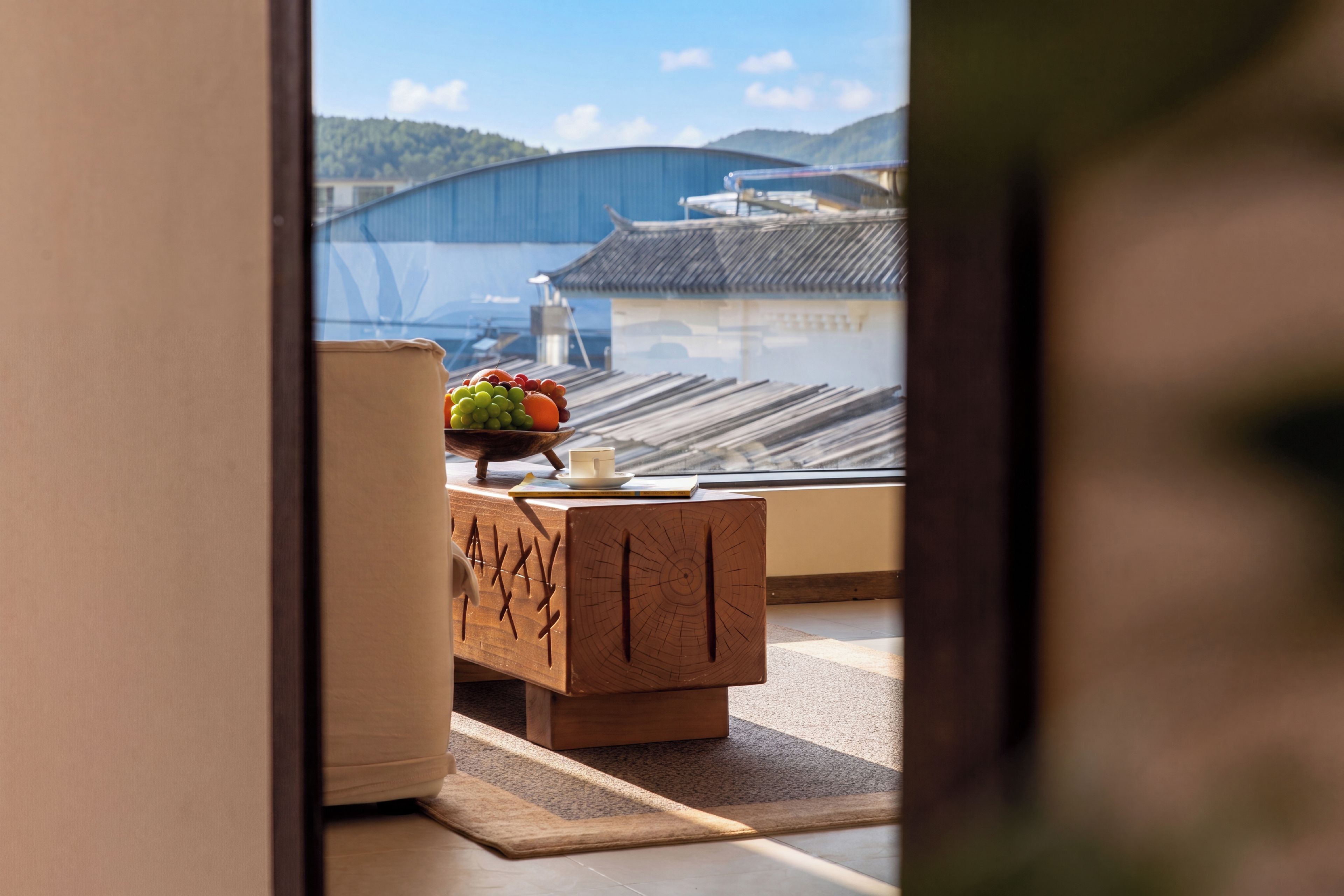 A room with a wooden table, a bowl of fruit, and a cup. Outside, there's a traditional building and a view of a hill.