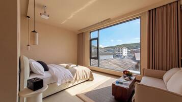 A hotel room with a bed, a desk, a chair, a large window with a view of buildings, and a projector mounted on the ceiling.