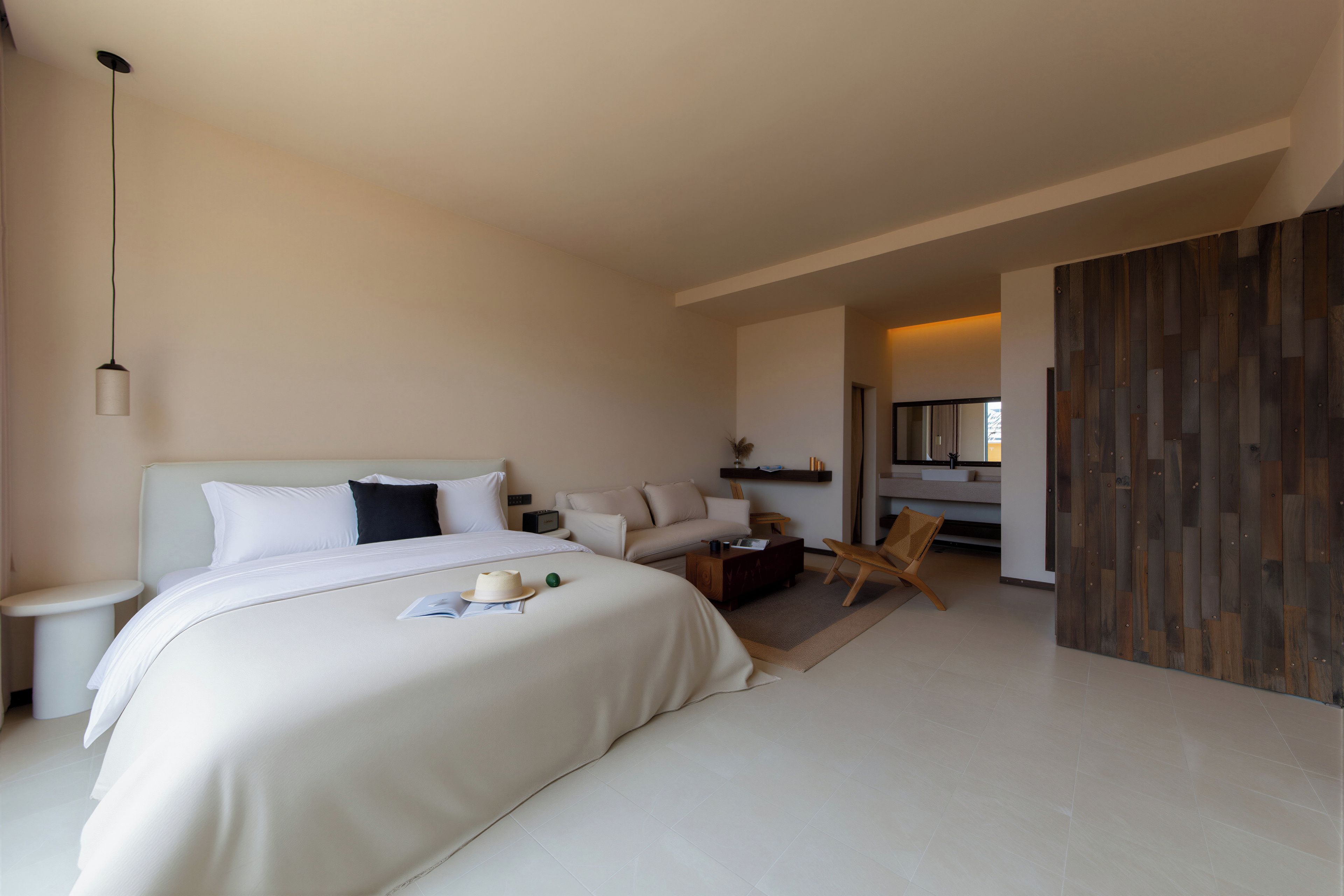 A modern hotel room with a large bed, a sitting area with a sofa and armchairs, a small table, and a wooden partition.
