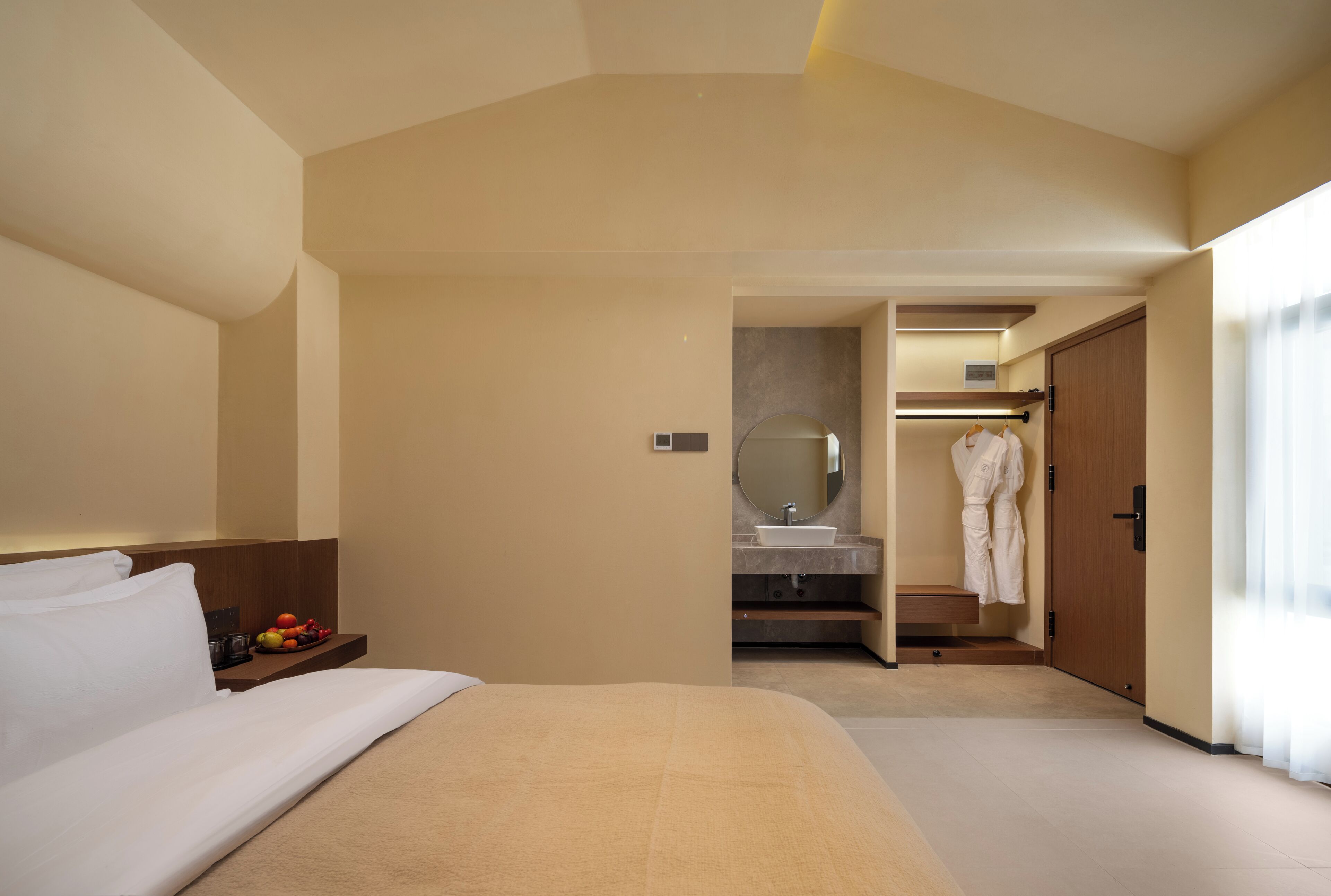 A hotel room with a bed, a bedside table with fruit, a bathroom with a sink and mirror, and a robe hanging on a door.
