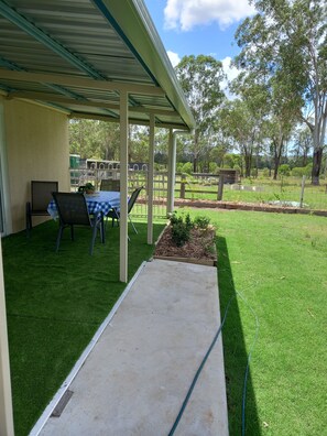 A covered patio area with a table and chairs, a concrete pathway, and a grassy lawn with trees in the background.