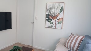 Een woonkamer met een flatscreen televisie aan de muur, een lijst met bloemenkunst, een bank met gestreepte kussens en een kleine plant op een houten tafel.