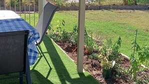 Een overdekt terras met een tafel en stoelen, omgeven door een verzorgd gazon en bloembedden.
