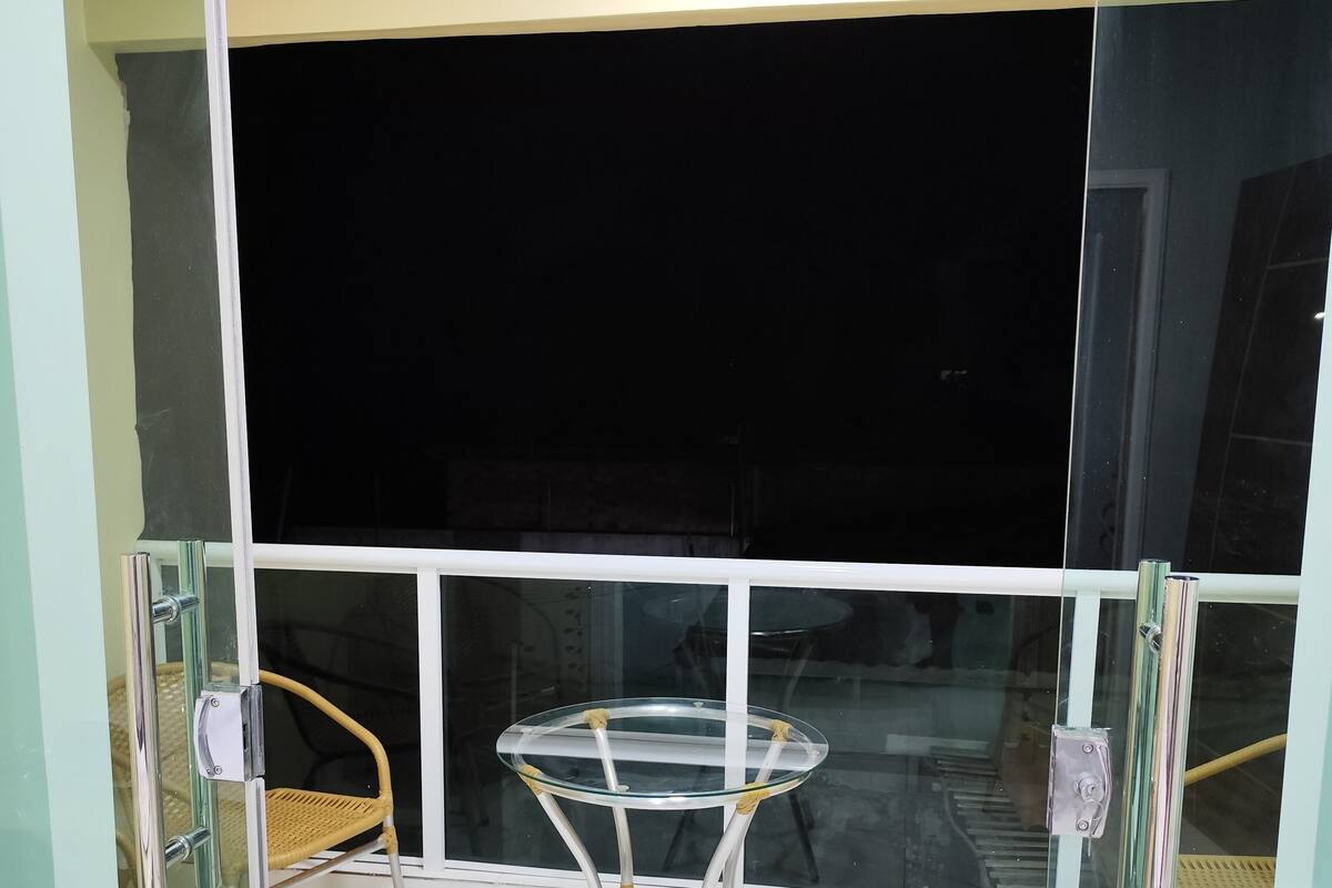 A balcony with a glass table and wicker chairs, a white tiled floor, and a large window.