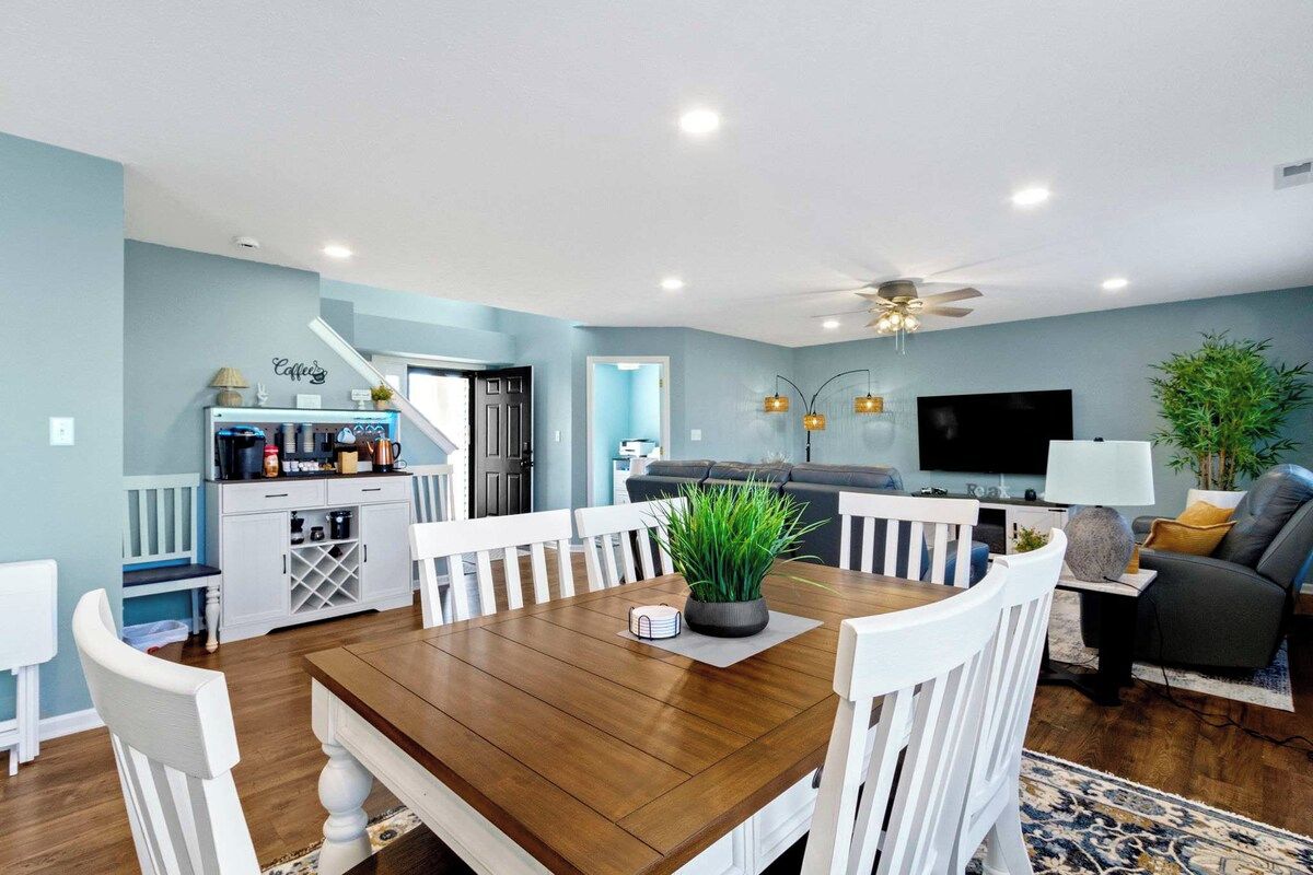A dining area with a wooden table and white chairs, a kitchen with a coffee station, a living room with a sofa and a TV, and a ceiling fan.