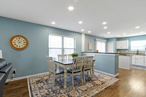 A dining area with a table and chairs, a clock on the wall, and a kitchen in the background.