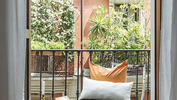 A balcony with a metal railing, a bed with pillows, and a view of a building with greenery.