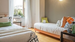A bedroom with two beds, a window with curtains, a bedside table with a lamp, and a view of buildings outside.