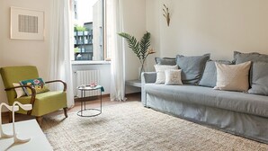 A living room with a green armchair, a grey sofa, a small side table, a large window with curtains, and a radiator.