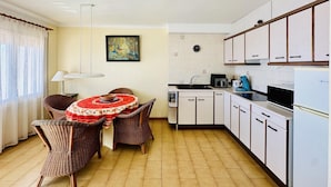 A compact kitchen with white cabinets, a dining area with wicker chairs, a red tablecloth, and a painting on the wall. - 2210 - MIRADOR II 55 WONDERFUL SEA VIEW, IDEAL SITUATION FOR HIKING, 150m. FROM THE BEACH OR FROM THE ROCKS. (Roses)